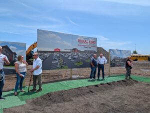 People stand on a strip of fake grass at a construction site in front of a rendering of the new Rural King Store Support Campus. The rendering depicts a parking lot in front of a red and white store.