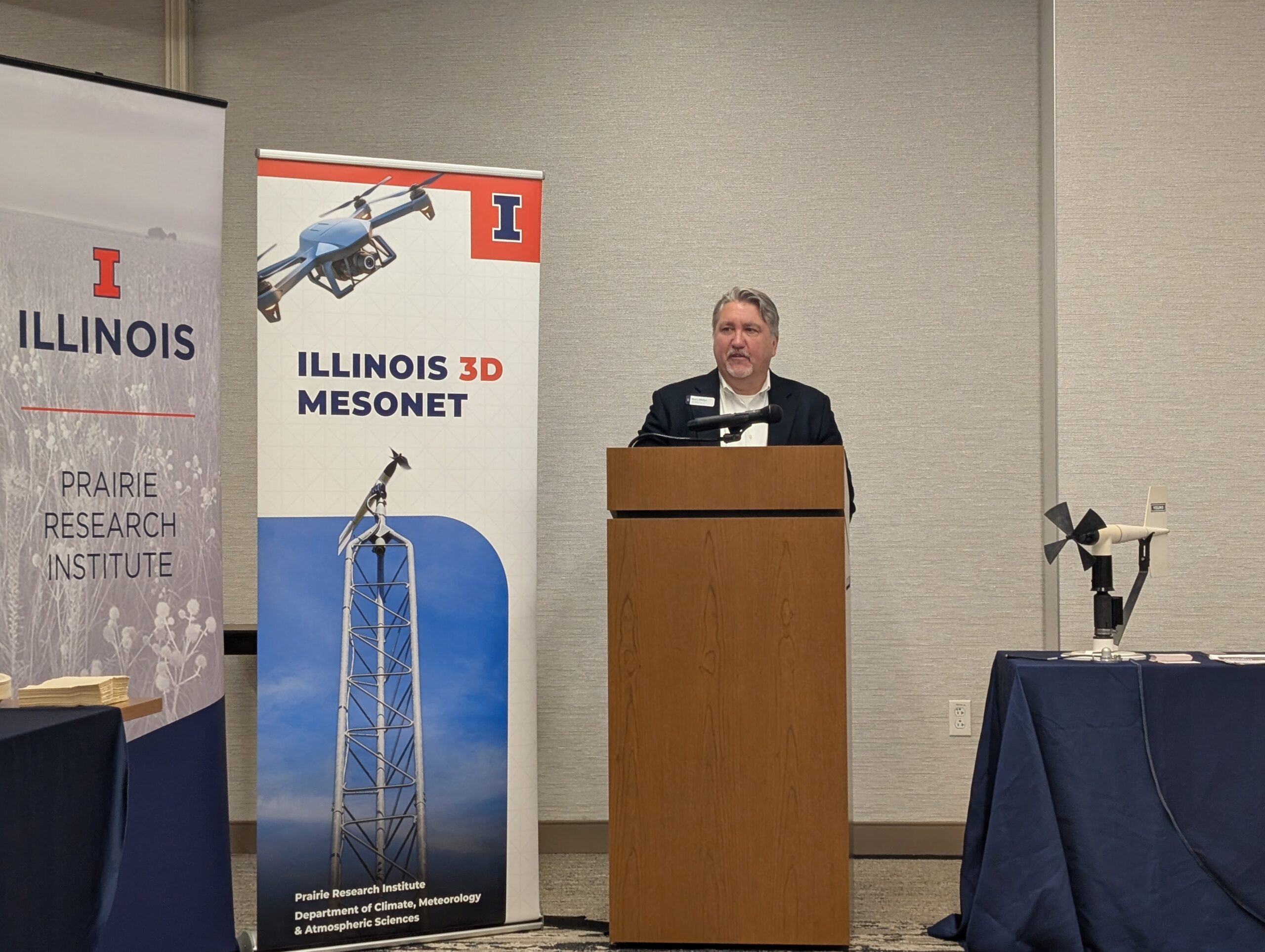 Prairie Research Institute Deputy Director Marc Miller stands behind a podium. To his left is a banner with a drone and a climate monitoring station. To his right is an anemometer.