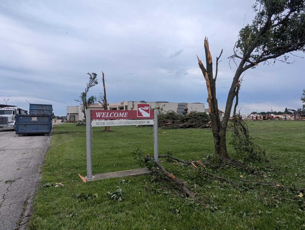 A welcome sign stands next to a damaged tree in front of a dumpster, a pile of branches and a flattened building.
