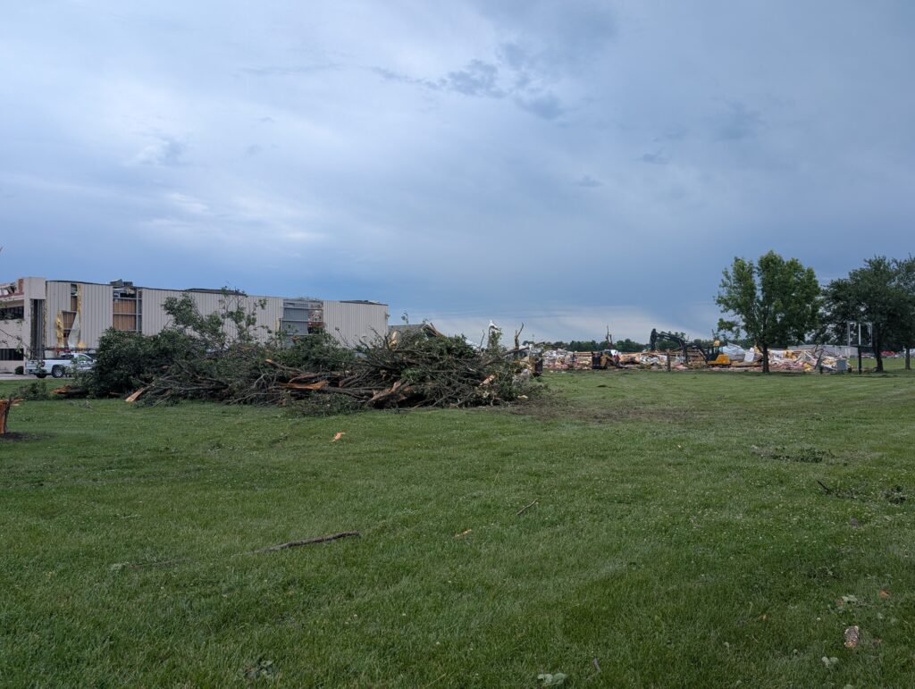 A pile of branches lays next to a damaged tree in front of a flattened building.