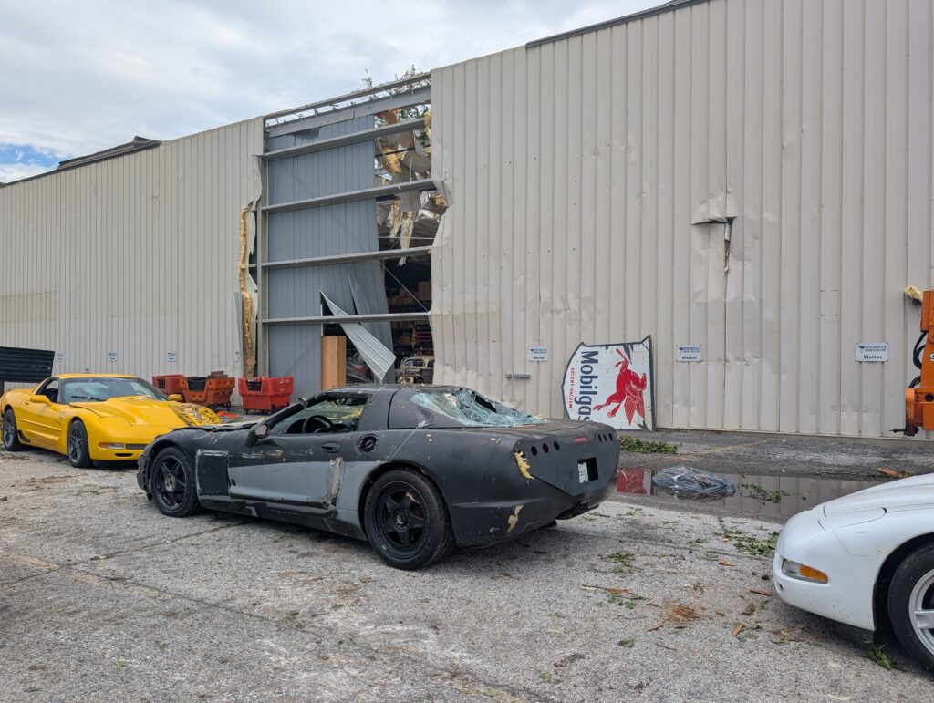 A smashed black and yellow Corvette sit outside of a damaged building.