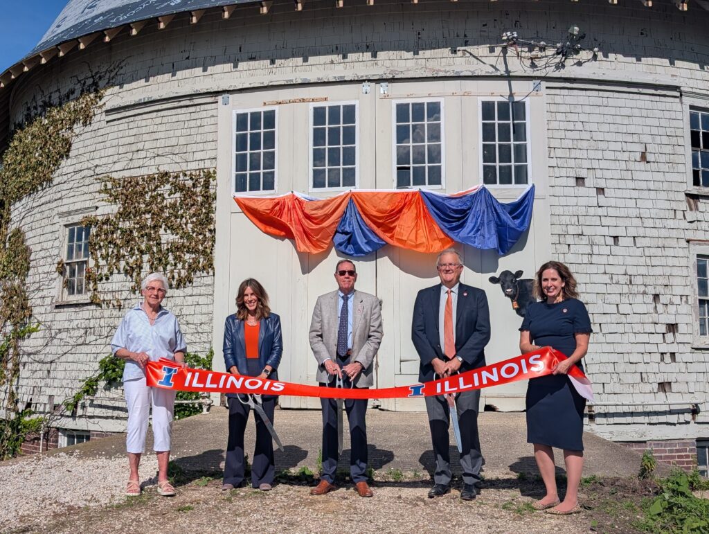 People stand behind an orange ribbon that says Illinois holding giant scissors in front of a white round barn with a cow decal on it.