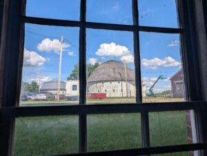 Looking through a window, there are two white round barns. Cars are parked to the left of them.