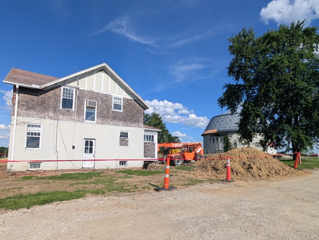 Orange cones and construction tape block off a house with an orange piece of construction equipment and a pile of dirt. A round barn and a tree are to the right.