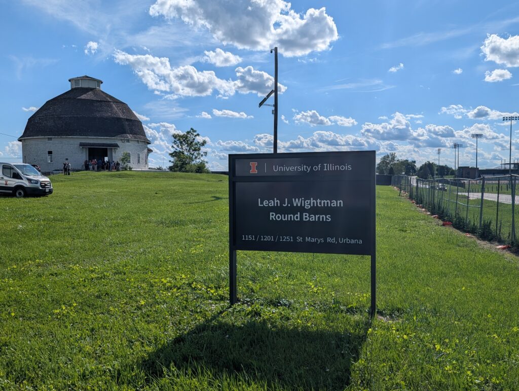 A blue sign reads, "University of Illinois Leah J. Wightman Round Barns." Behind it, stands a white round barn.