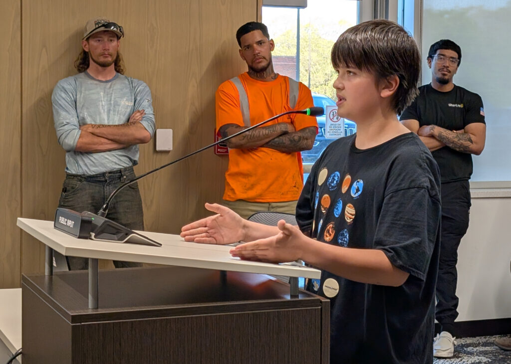 A boy speaks into a microphone behind a podium while gesturing with his hands. 