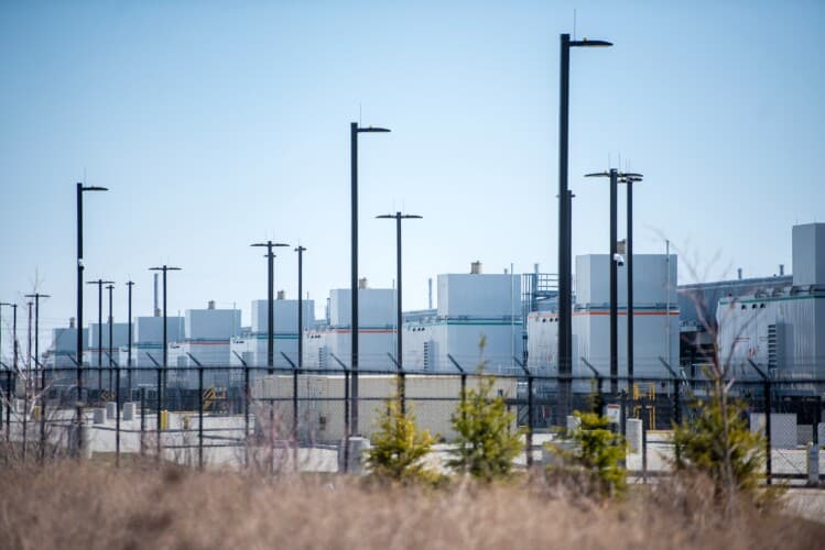 A line of buildings sit behind a fence and street lights. 
