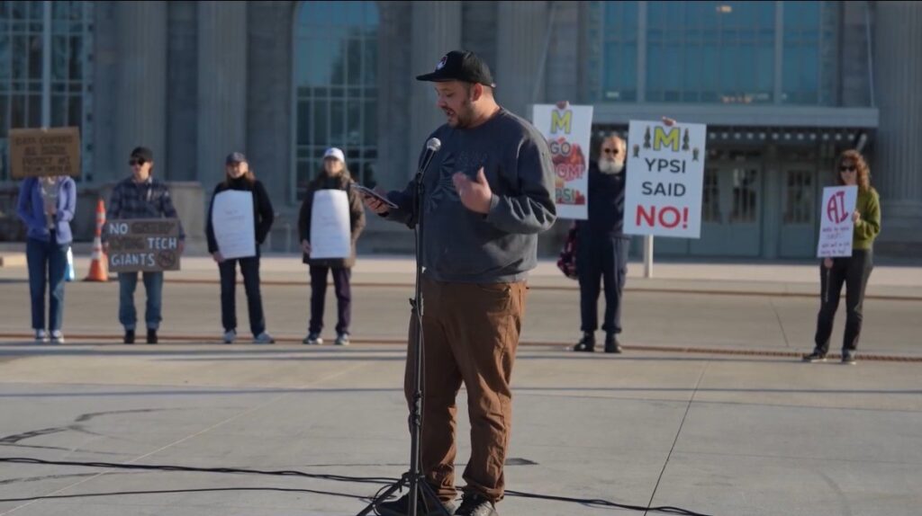 Michigan State Rep. Dylan Wegela speaks into a microphone in front of a line of people holding signs.