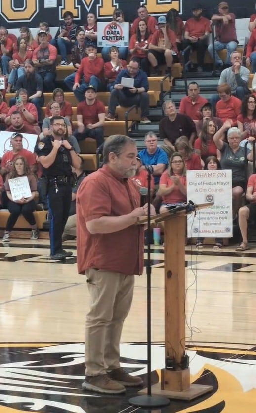 A man speaks into a microphone behind a podium in a gymnasium. People sit in bleachers behind him holding signs.