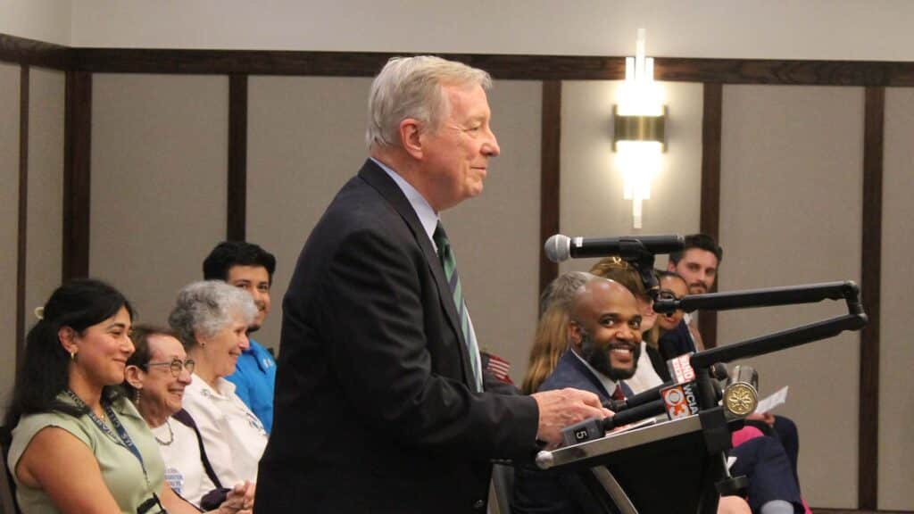 An elderly man in a suit speaks at a podium while other officials present laugh.
