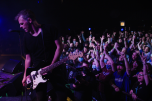 A man in a black T-shirt plays guitar in front of a crowd of people raising their arms up.