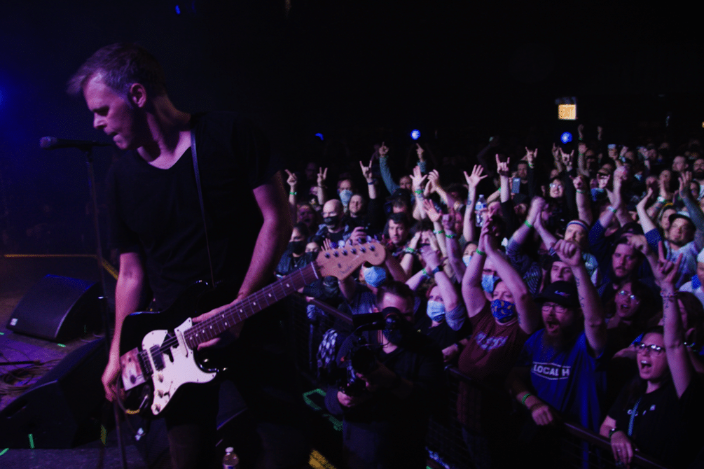 A man in a black T-shirt plays guitar in front of a crowd of people raising their arms up.