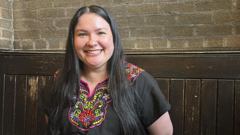 A woman with dark hair wearing a black blouse with a colorful pattern embroidered around the collar.