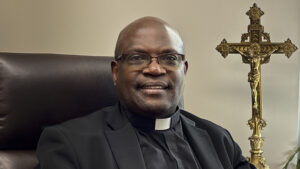 An older black man wearing glasses and priest's attire sits in an office chair in front of a golden crucifix.