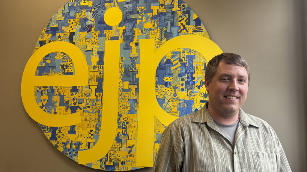 A white man in a light button-up shirt stands and smiles to the camera in front of a large, circular sign with a blue and yellow painted pattern and three large lowercase yellow letters reading "ejp."