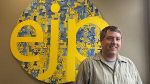 A white man in a light button-up shirt stands and smiles to the camera in front of a large, circular sign with a blue and yellow painted pattern and three large lowercase yellow letters reading "ejp."