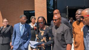 State Rep. Carol Ammons reads out of a notebook in front of a brick building. Her lawyer, Mataqee Akbar, stands to her right, and her husband, Champaign County Clerk Aaron Ammons, stands to her right. They are surrounded by other supporters.
