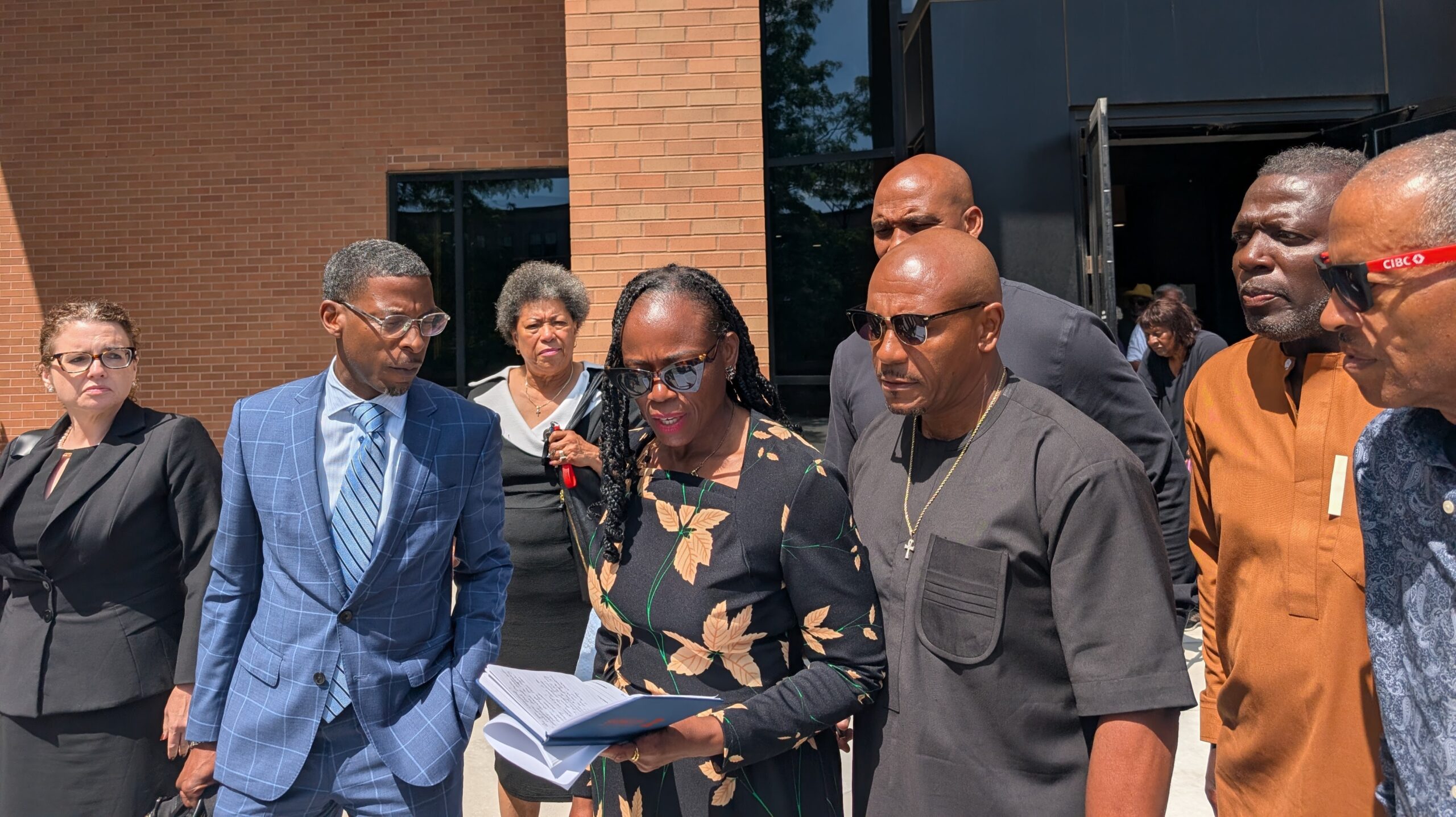 State Rep. Carol Ammons reads out of a notebook in front of a brick building. Her lawyer, Mataqee Akbar, stands to her right, and her husband, Champaign County Clerk Aaron Ammons, stands to her right. They are surrounded by other supporters.