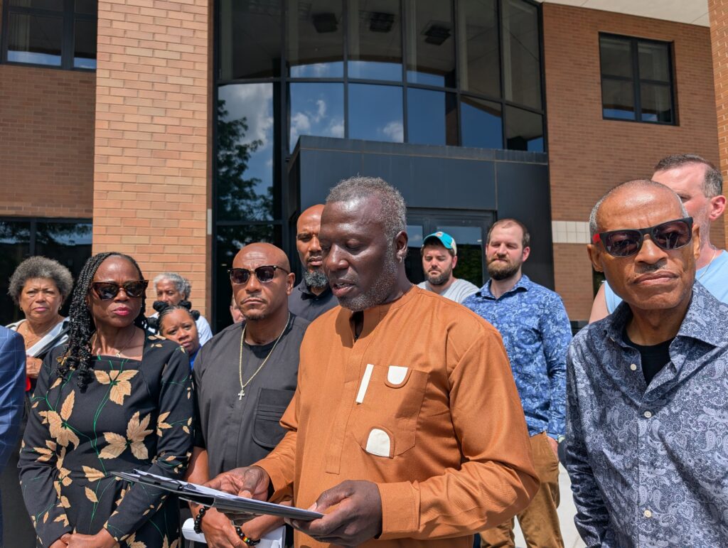 Reparations United Director Kamm Howard reads off a piece of paper on a clipboard in front of a brick building. State Rep. Carol Ammons and Champaign County Clerk Aaron Ammons stand to his left. Other supporters surround him. 