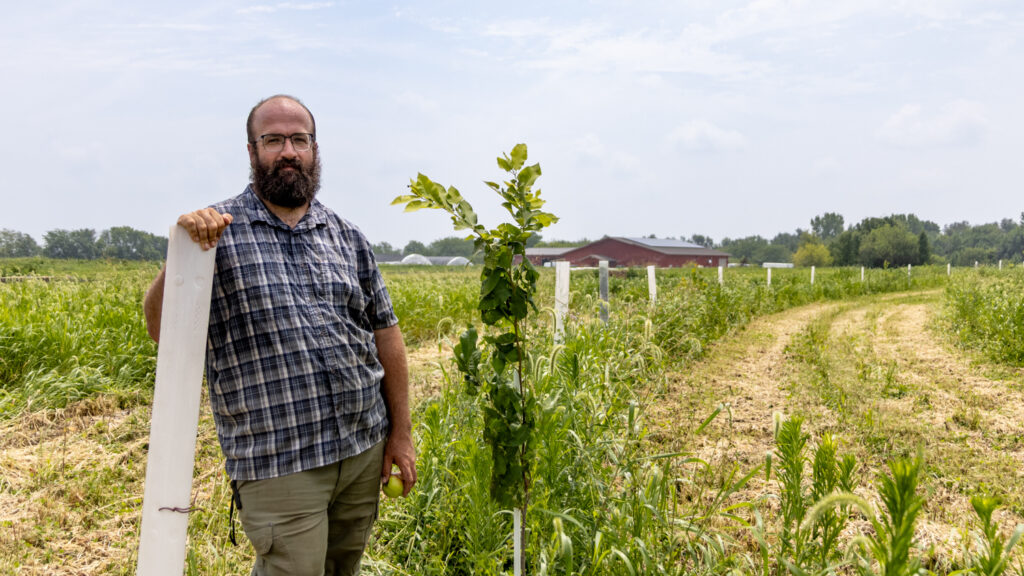 A man with a brown beard wearing glasses, a blue plaid shirt and green pants leans against a white tube. The white tubes run into the background and are surrounded by fields.