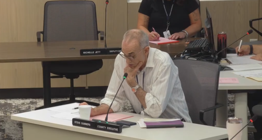 Champaign County Executive Steve Summers leans forward with his chin in his hand as he speaks into a microphone.