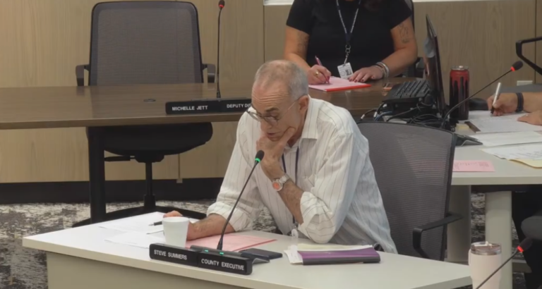 Champaign County Executive Steve Summers leans forward with his chin in his hand as he speaks into a microphone.