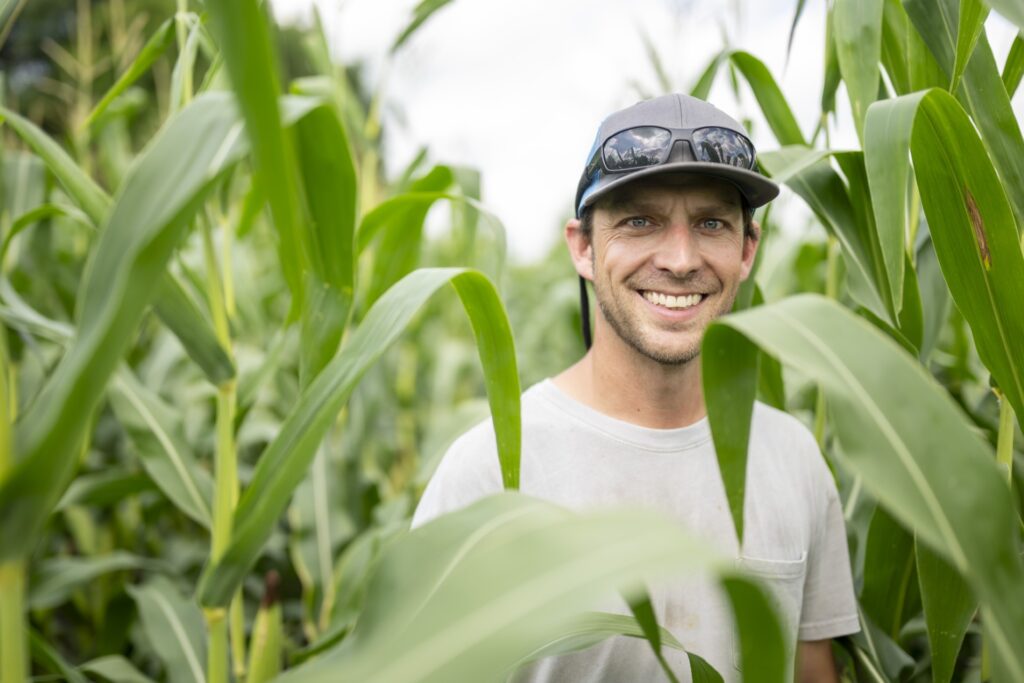 A man wearing a gray baseball hat with sunglasses perched on top wearing a white t-shirt smiles for a picture in a field of corn.