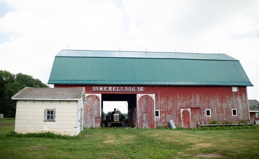 A tractor sits in a faded red barn. A tan shed sits in front of it to the left.