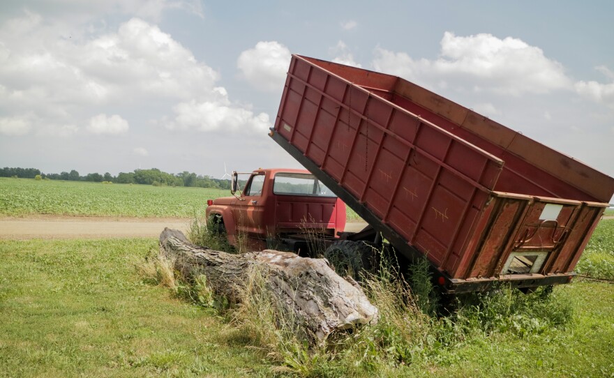 A red truck with a red bed tilting up faces a dirt road. A log sits to the left of it.