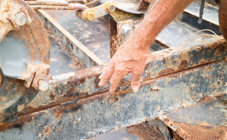 A hand reaches down to touch a piece of rusting farm equipment.