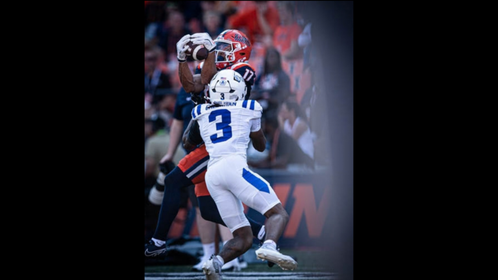 An Illinois football player catches the ball in the air ahead of a blue and white Duke player.