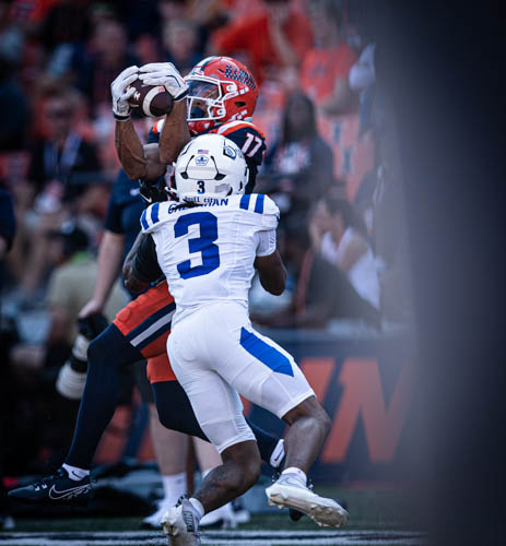An Illinois football player catches the ball in the air ahead of a blue and white Duke player.