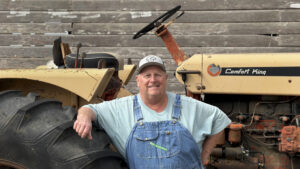 A white man in a blue T-shirt and overalls smiles and leans on the large back wheel of an antique, yellow tractor in front of a faded wooden wall.