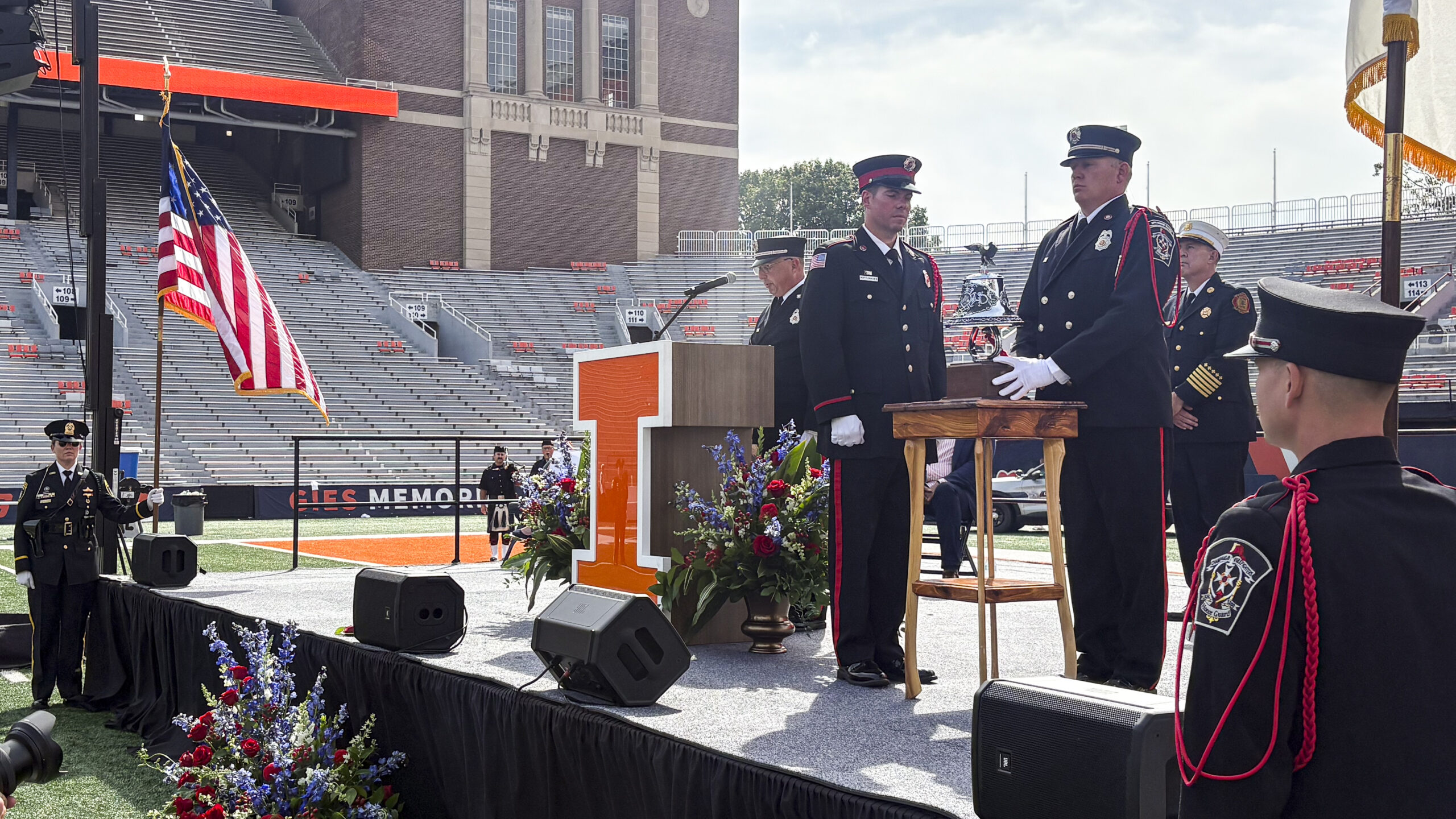 Men dressed in formal firefighter attire stand on a stage with a large orange letter I podium and an American flag and ring a silver bell. The stage they stand on is situated on a football field within a large stadium and morning sunlight streams past the clouds into the stadium.
