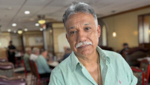 An older man in a light green shirt with grey-white hair and a grey-white mustache looks into the camera. The background is a warm, well-lit restaurant interior dining room.