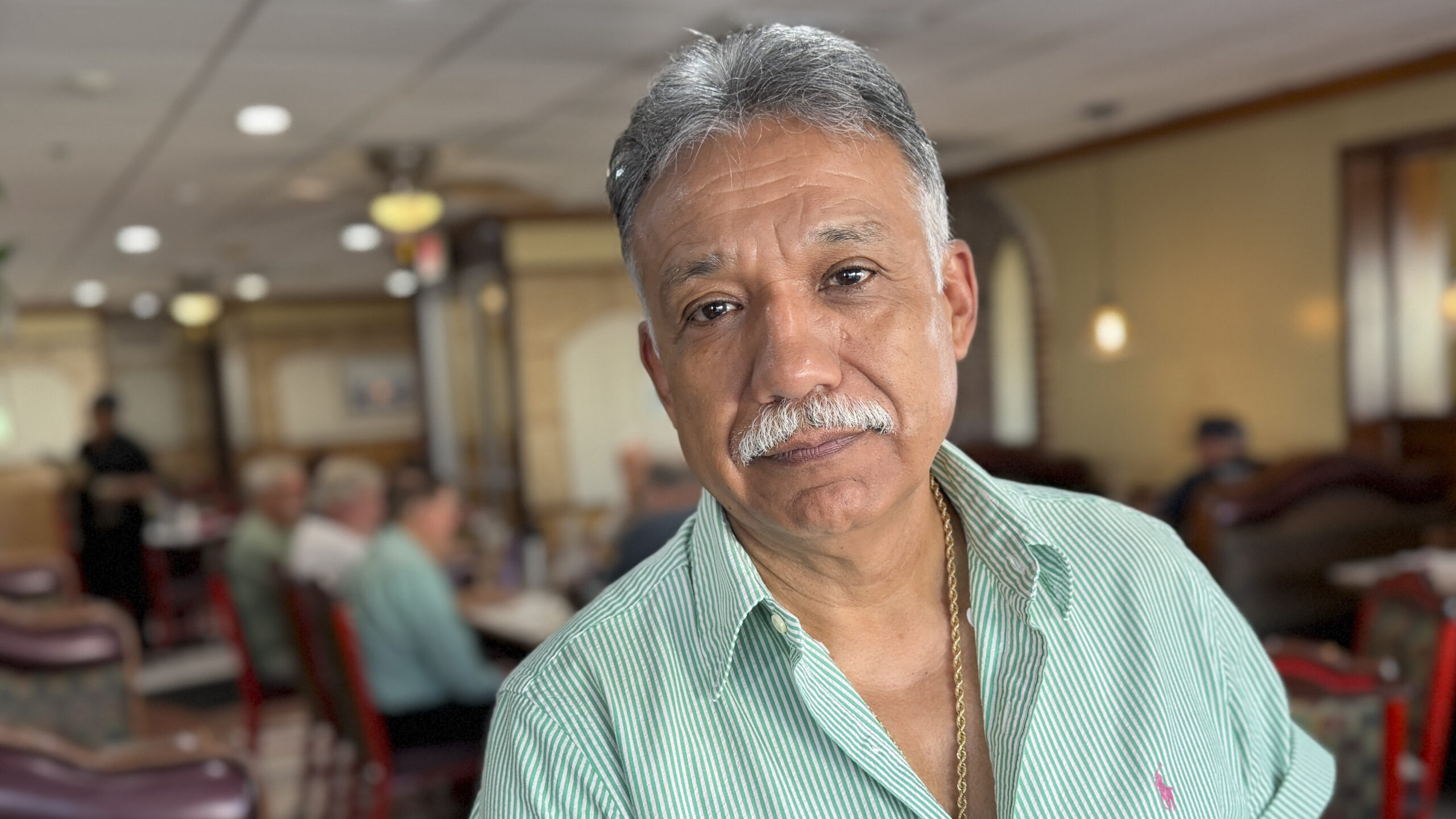 An older man in a light green shirt with grey-white hair and a grey-white mustache looks into the camera. The background is a warm, well-lit restaurant interior dining room.
