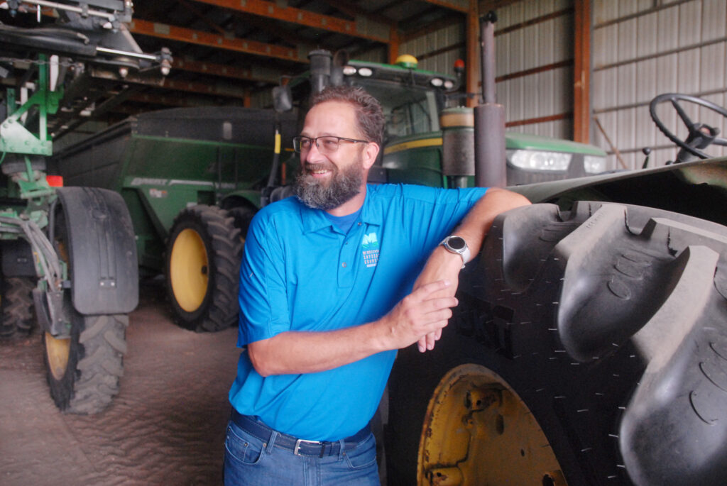A man wearing glasses. a blue polo shirt and jeans leans against the tire of a green tractor in a barn.