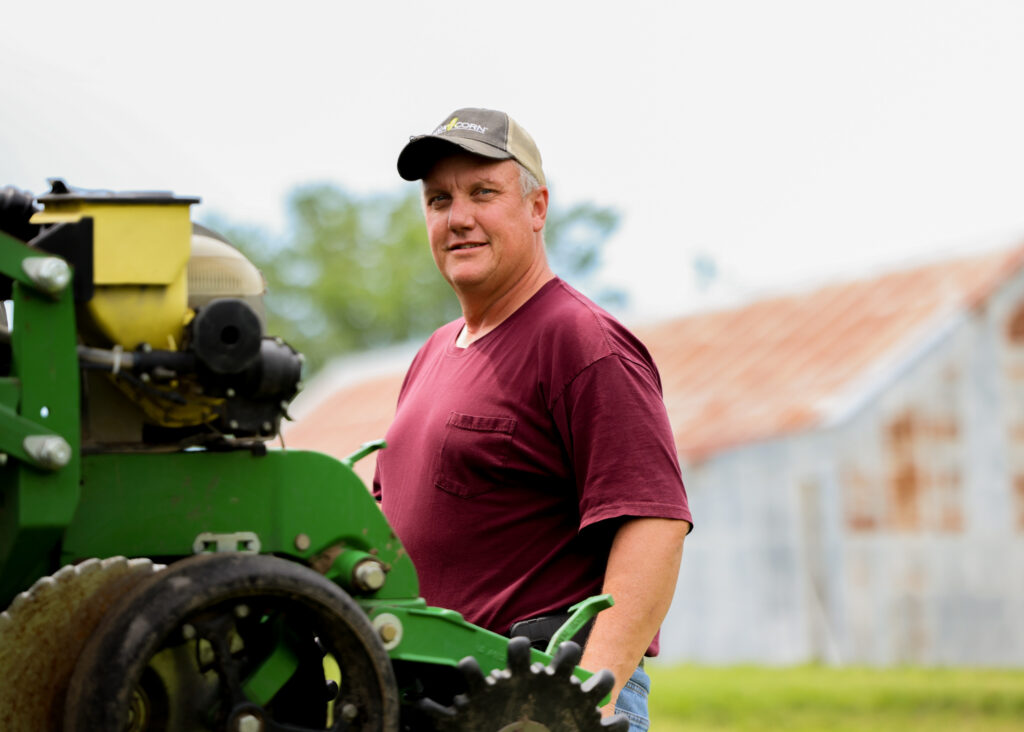 A man wearing a baseball hat, a maroon t-shirt and jeans stands behind a green piece of farm equipment. 
