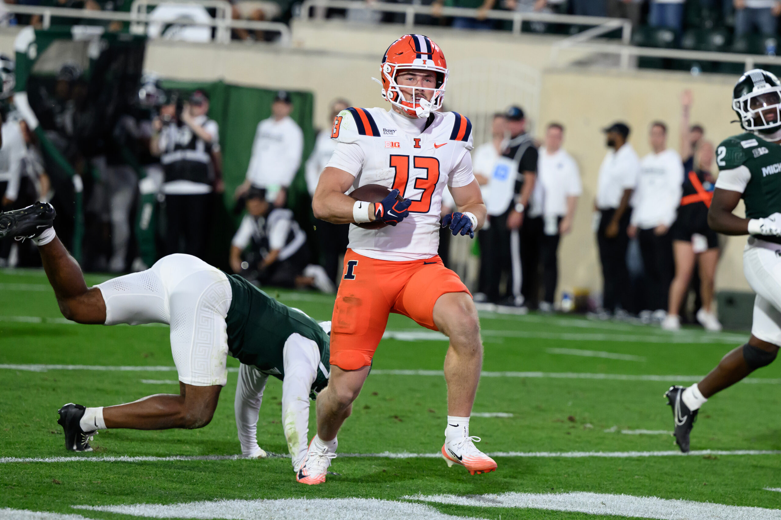 An Illini football player wearing orange and white holds a football and runs down the field. An MSU player falls to the ground behind him.