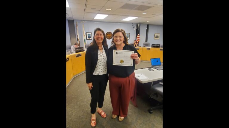 Two women stand next to each other. The one on the right holds up a certificate with the oath of office for a new Urbana city council member.