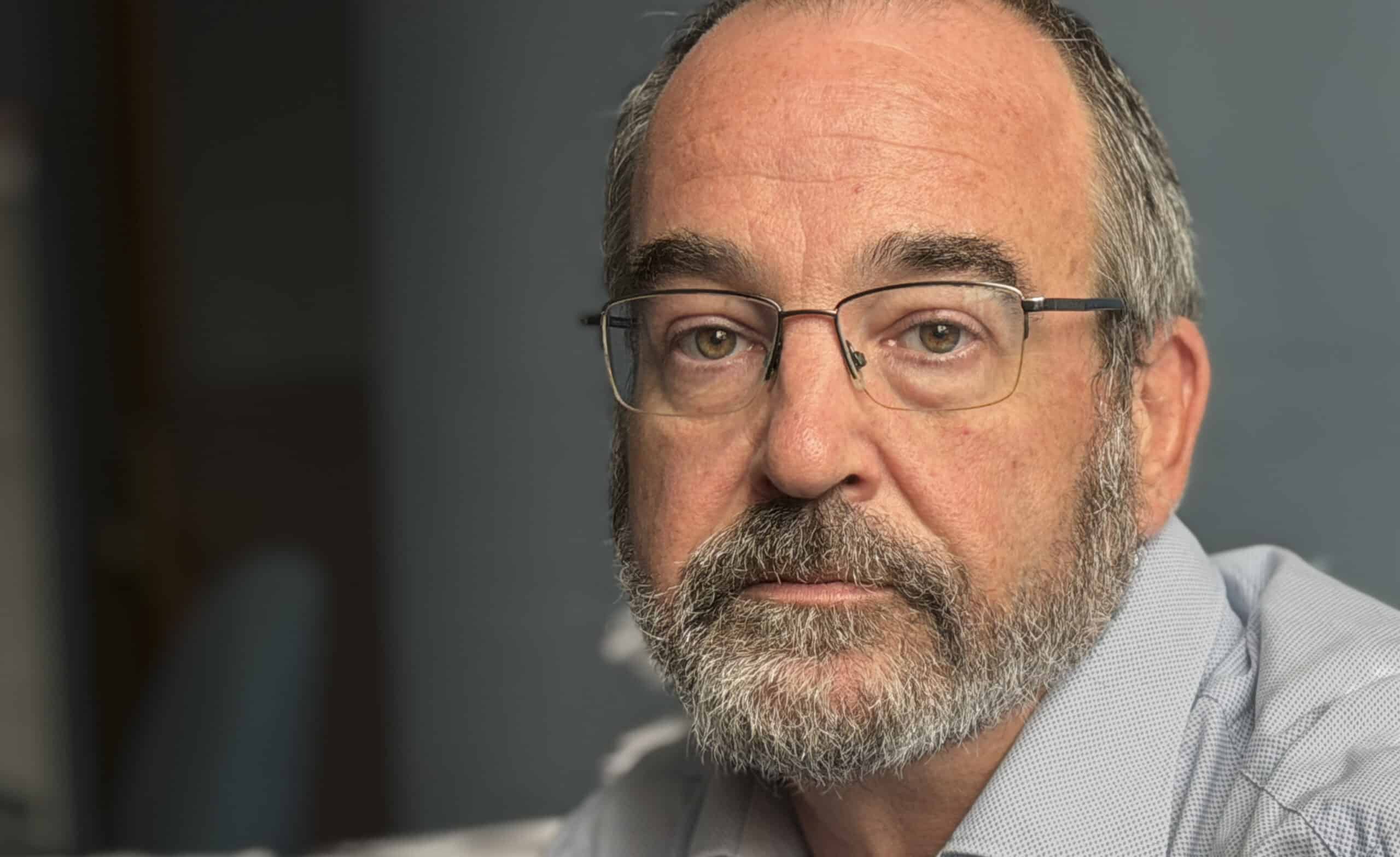 An older white man wearing a collared shirt with gray hair and glasses looks toward the camera.