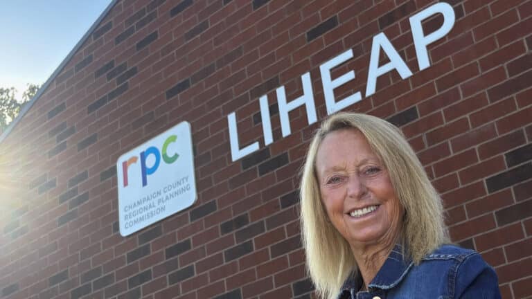 A white woman with blonde hair wearing a blue polo shirt stands in front of a brick building with a white logo reading "LIHEAP" and smiles at the camera.