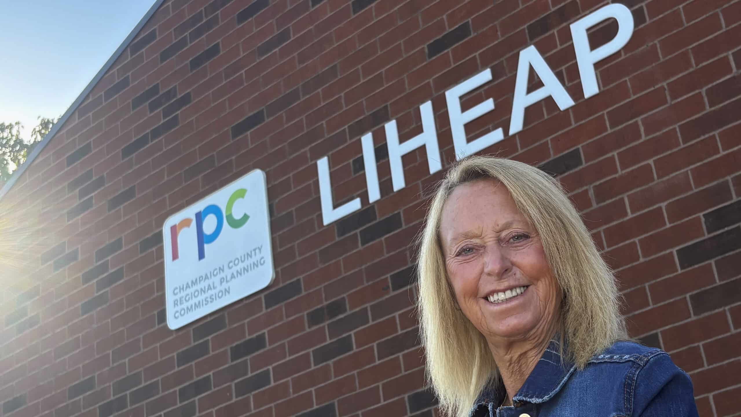 A white woman with blonde hair wearing a blue polo shirt stands in front of a brick building with a white logo reading "LIHEAP" and smiles at the camera.