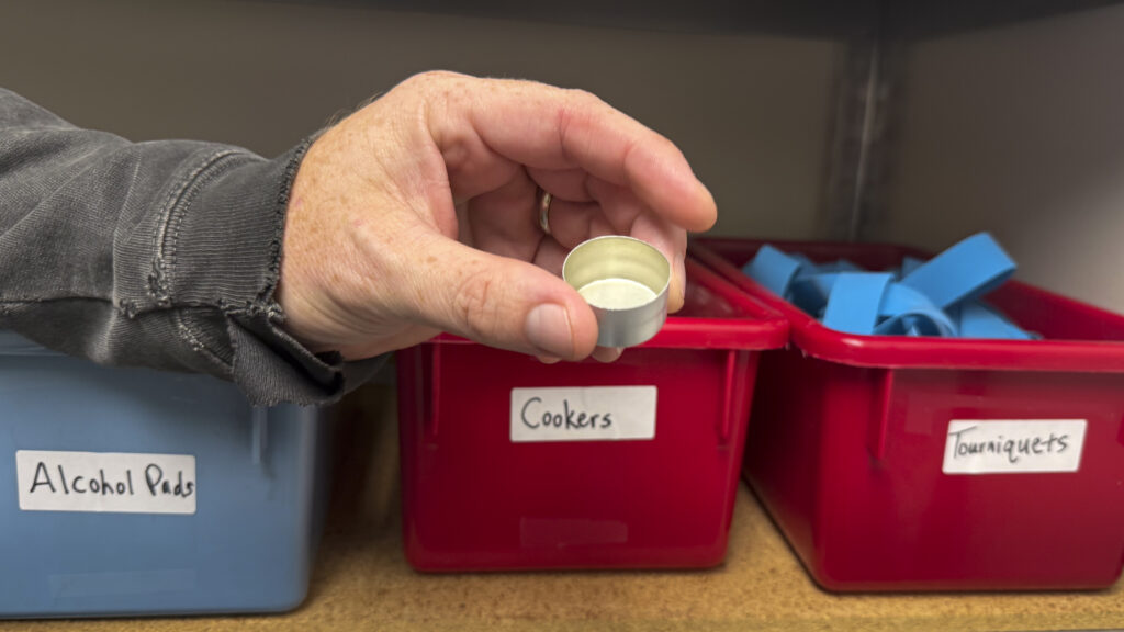 a person holds a small metal cup. boxes on a shelf are labeled "alcohol pads," "cookers" and "tourniquets"