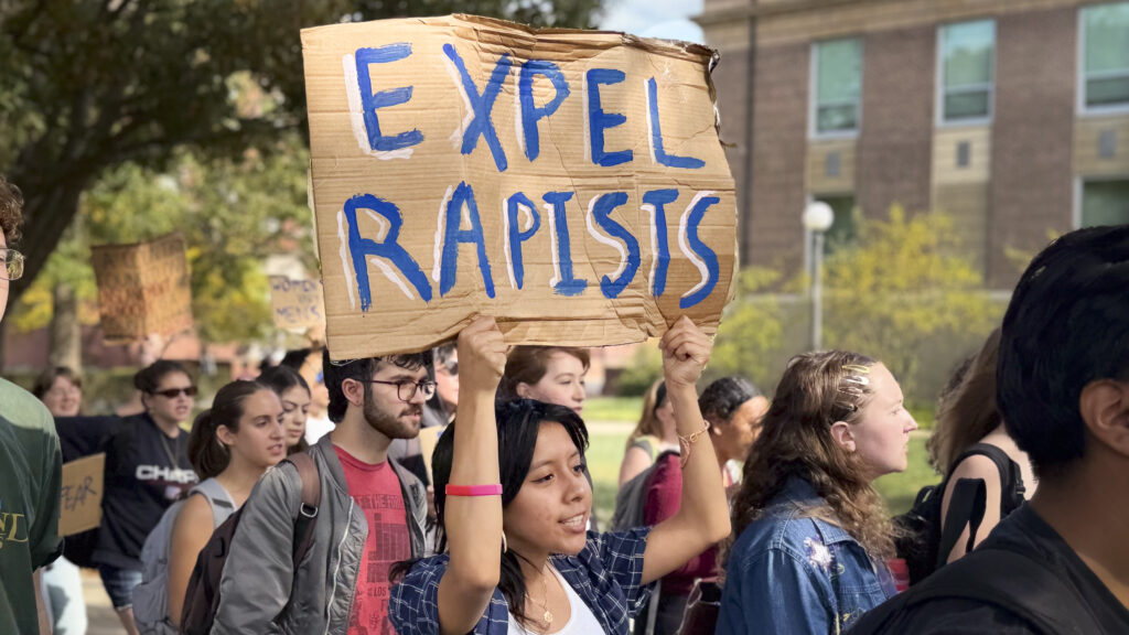attendees at a rally outside. one person holds a sign that reads: Expel Rapists