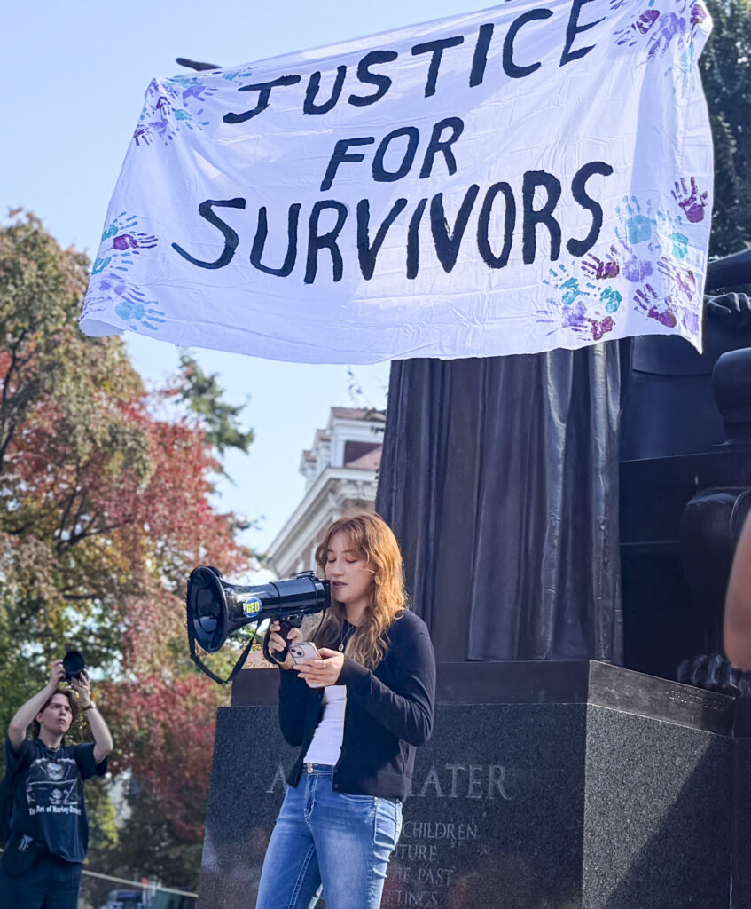 A person stands outside and speaks into a megaphone. A sign over her head says "Justice for Survivors"