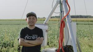 A man smiles at the camera while standing in front of a tall machine that stretches out of the top of the frame and a field of green crops. He wears a black T-shirt and a green baseball cap.