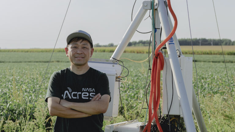 A man smiles at the camera while standing in front of a tall machine that stretches out of the top of the frame and a field of green crops. He wears a black T-shirt and a green baseball cap.