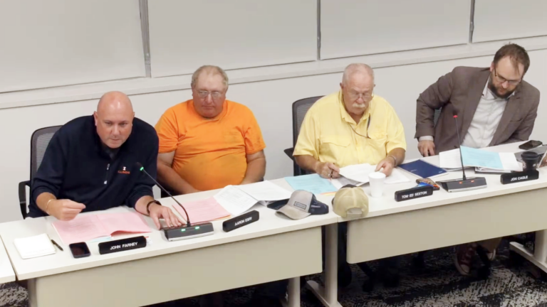 A screenshot of four white men sitting along a table at a county board meeting.