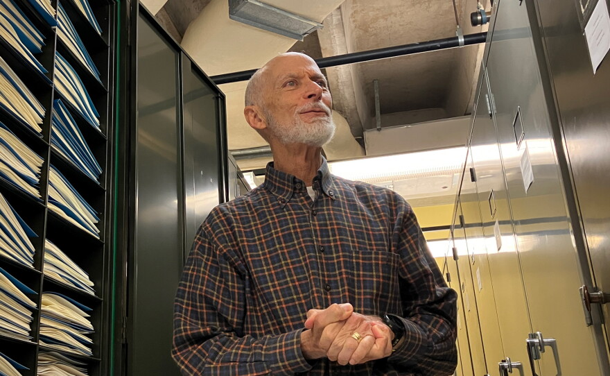 A man clasps his hands standing between two lines of cabinets.